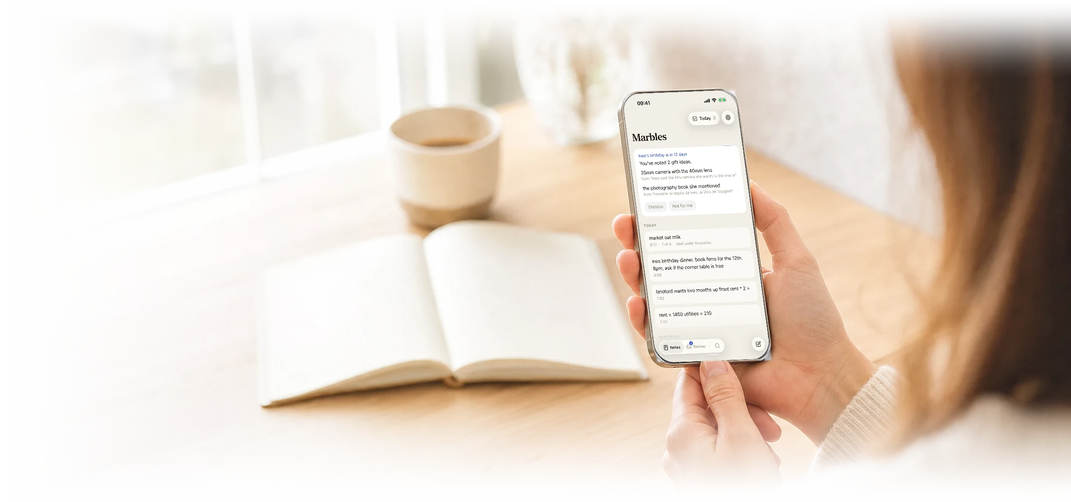 A person at a table by a bright window holding an iPhone. Marbles is showing a card about a birthday coming up and the gift ideas it found, above the day’s notes. An open notebook and a coffee sit beside them.
