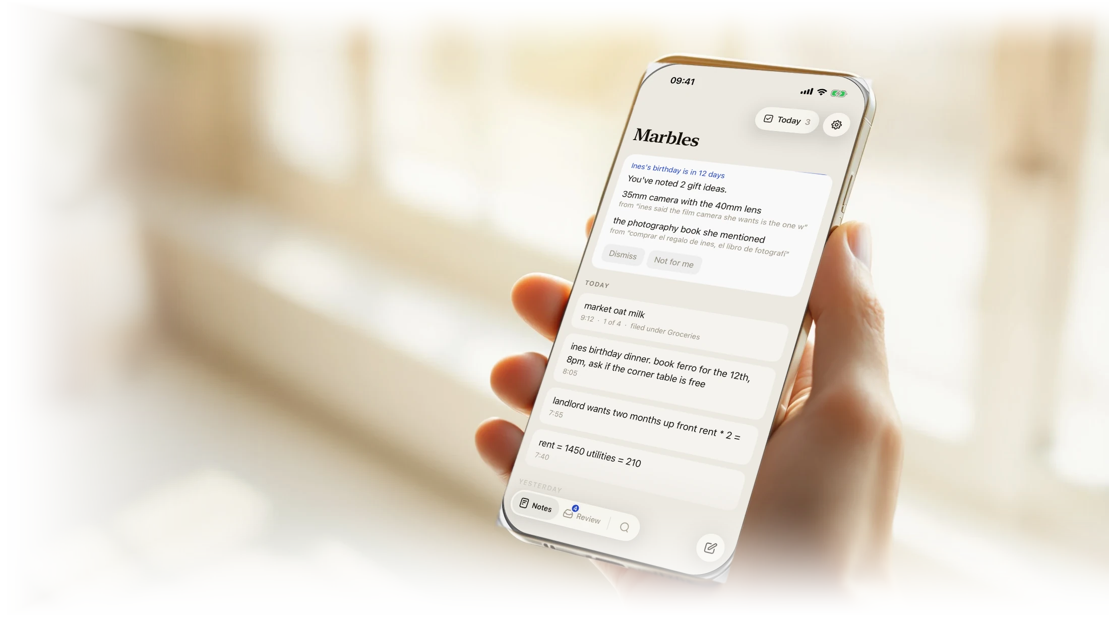 A person holding an iPhone by a sunlit window, Marbles on the screen showing a birthday reminder with the gift ideas it found, above the day’s notes.
